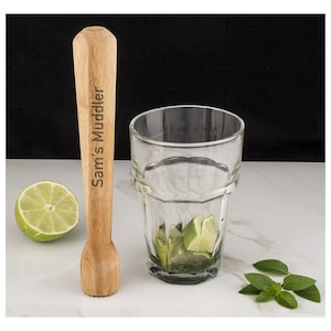 May include: A wooden muddler with the text "Sam's Muddler" engraved on it. The muddler is next to a glass with lime wedges and mint leaves.
