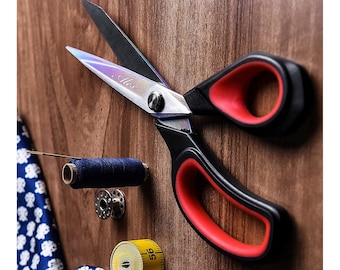 May include: A pair of black and red fabric shears with a silver blade. The shears are on a wooden surface with a spool of blue thread, a sewing needle, and a yellow measuring tape.