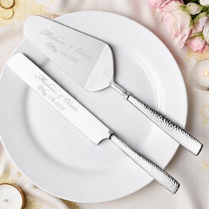 May include: A silver cake server and knife set on a white plate. The set is engraved with "Michael & Olivia" and "May 15th 2025". The handles have a textured design, and the set is likely for a wedding or special occasion.