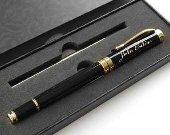 May include: A black pen with gold accents and the name "John Collins" engraved on the barrel, presented in a black gift box. The pen features a gold clip and trim, and the box has a decorative pattern on the lid.