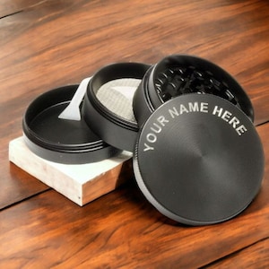 May include: Black metal grinder with a removable lid and a compartment for storing herbs. The grinder has a personalized inscription that reads "YOUR NAME HERE".