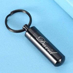 Puede incluir: Un llavero cilíndrico negro con la inscripción "David" en color plata. Está unido a un llavero negro. El fondo es de color azul claro.