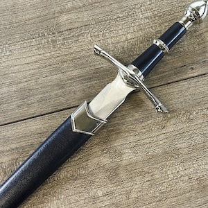 May include: A silver and black dagger with a black leather handle and a silver pommel. The dagger is on a wooden surface.