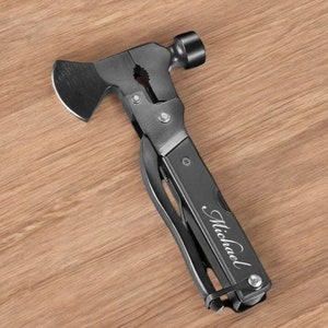 May include: A multi-tool with a hammer, axe, and pliers. The tool is dark gray with the name "Michael" engraved on the side. The tool is lying on a wooden surface.