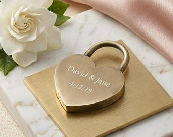 May include: A heart-shaped brass padlock engraved with "David & Jane" and "4/12/18" rests on a gold square. A white gardenia flower with green leaves sits on a marble and gold surface, with a pink satin fabric in the background.