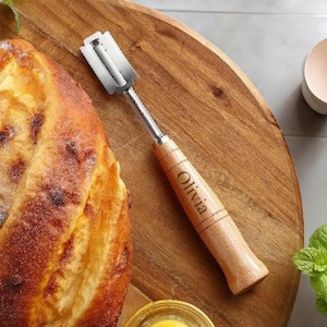 May include: A wooden bread lame with a silver blade, engraved with the name "Olivia". The lame is on a wooden surface next to a loaf of bread.