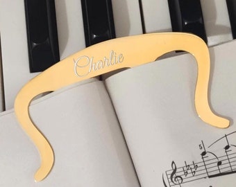 May include: A gold-colored page holder with the name "Charlie" engraved on it. The page holder is resting on a sheet of music with musical notes.