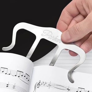 May include: A silver music page holder with the word "Parker" engraved on it. The holder is designed to hold open a book of sheet music.