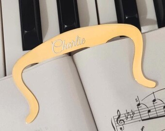 Peut inclure: Porte-pages en métal doré avec le nom "Charlie" gravé dessus. Le porte-pages est posé sur une partition de musique sur un clavier de piano.