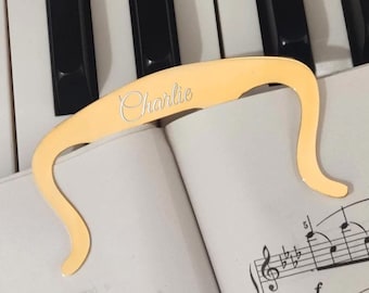 Peut inclure: Un serre-page doré avec le nom "Charlie" gravé, posé sur une partition musicale. Le support a un design incurvé et est placé sur un clavier de piano.