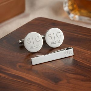 May include: Two circular silver cufflinks and a matching silver tie bar. Each cufflink is engraved with the initials S|C and the date 10.3.23. The tie bar features the same S|C engraving on one end, all presented with a brushed metallic finish.