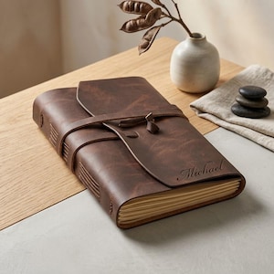 May include: A dark brown leather journal with a wrap-around strap closure and visible stitching along the spine. The front cover features the name Michael engraved in a cursive script. The journal contains cream-colored pages with deckled edges.