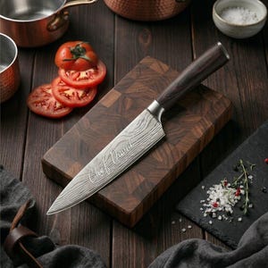 May include: A chef's knife with a patterned blade and a dark wooden handle rests on a wooden cutting board. The blade is engraved with the words "Chef David". Sliced tomatoes and a small bowl of salt are also visible.