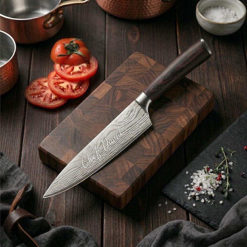 May include: A chef's knife with a patterned blade and a dark wooden handle rests on a wooden cutting board. The blade is engraved with the words "Chef David". Sliced tomatoes and a small bowl of salt are also visible.