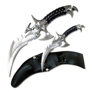 May include: Two silver and black fantasy style daggers with ornate designs and black leather handles. The daggers are in a black leather sheath.