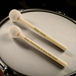 May include: Two wooden drumsticks with white felt heads rest on a drum. One stick is inscribed with "Aidan Jones," and the other reads "Keep drumming to your own beat!" The drum has a silver rim and a light gray surface.