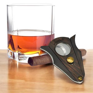 Puede incluir: Un cortapuros de madera oscura con una cuchilla circular de metal, un pomo de lat&oacute;n y el nombre James grabado en la superficie. El cortapuros est&aacute; colocado junto a un cigarro marr&oacute;n y un vaso transparente con un l&iacute;quido de color &aacute;mbar.