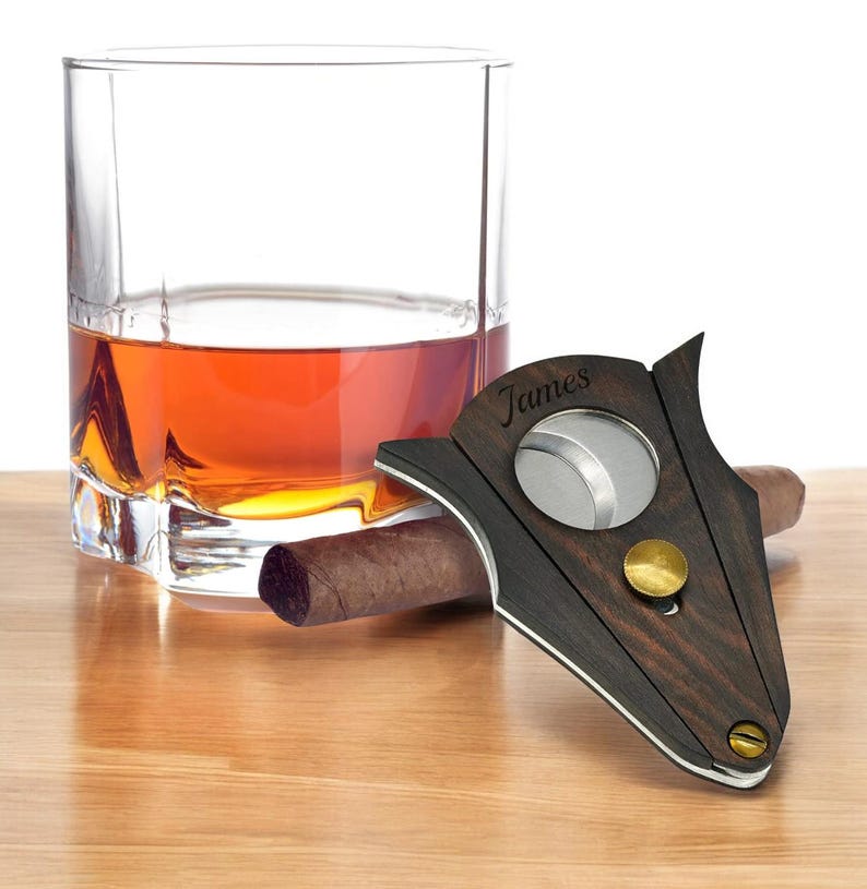 Puede incluir: Un cortapuros de madera oscura con una cuchilla circular de metal, un pomo de lat&oacute;n y el nombre James grabado en la superficie. El cortapuros est&aacute; colocado junto a un cigarro marr&oacute;n y un vaso transparente con un l&iacute;quido de color &aacute;mbar.