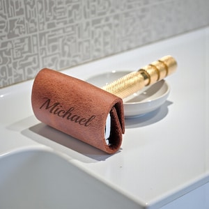 May include: A gold-colored safety razor with a textured handle rests in a small white dish. A brown leather razor cover with the name "Michael" engraved on it is next to the razor. The razor and cover are on a white surface.