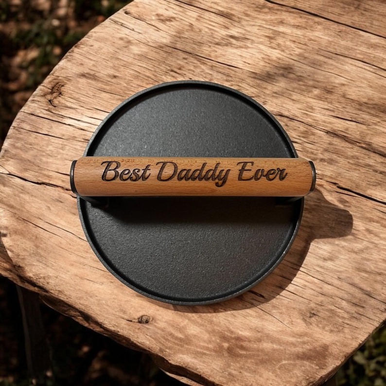 May include: A round, black cast iron burger press with a wooden handle. The handle is engraved with the words "Best Daddy Ever". The press is sitting on a rustic wooden surface.