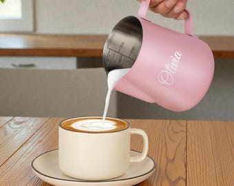 May include: A pink stainless steel milk frothing pitcher with the name "Olivia" printed on the side, pouring milk into a white coffee cup with a saucer. The cup has a brown rim and a latte art design.