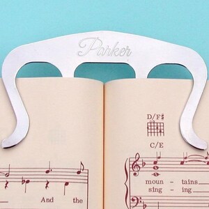 May include: Silver metal page holder with the word "Parker" engraved on it. The page holder is holding open a book of sheet music with musical notes and the words "And the" and "moun - tains. sing - ing."