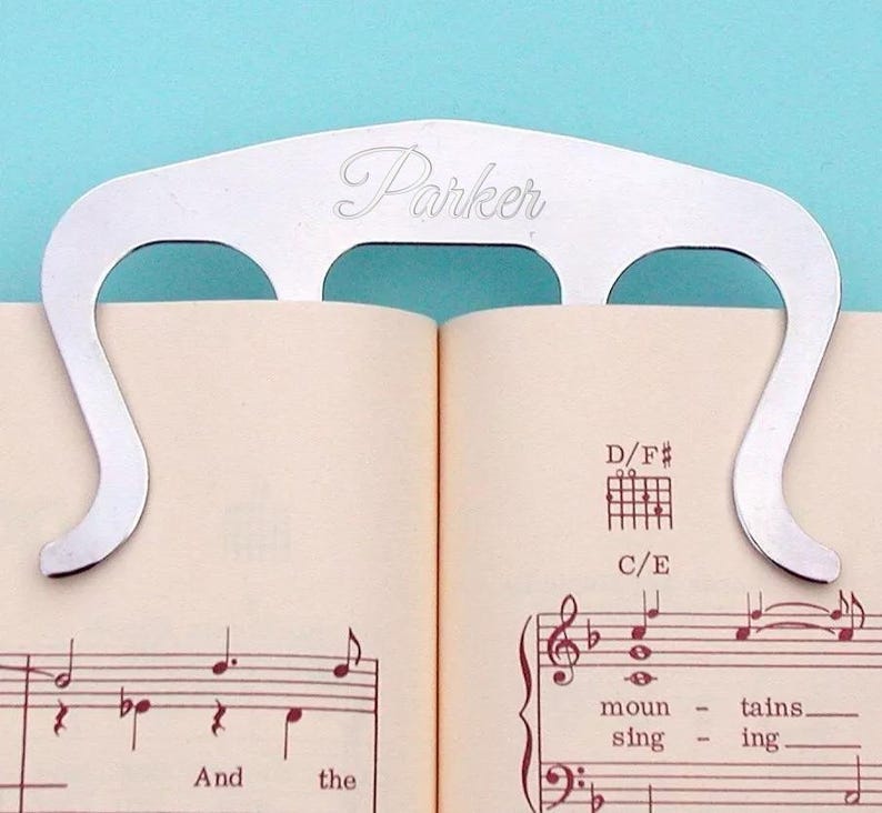 May include: Silver metal page holder with the word "Parker" engraved on it. The page holder is holding open a book of sheet music with musical notes and the words "And the" and "moun - tains. sing - ing."