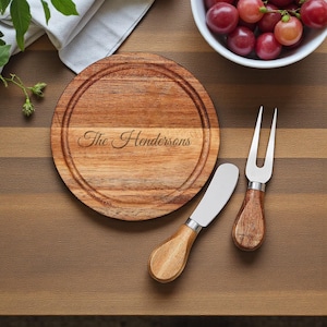 May include: A wooden cheese board with the engraved name "The Hendersons" is accompanied by a cheese knife and fork. The board has a circular groove and is made of light brown wood. The knife and fork have wooden handles and silver blades.