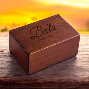 May include: A dark brown wooden box with the name "Bella" engraved on the top, along with the dates "2012-2025".