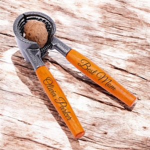 May include: A silver nutcracker with wooden handles. The handles are engraved with the text "Best Mom" and "Olivia Parker". A walnut is in the nutcracker.