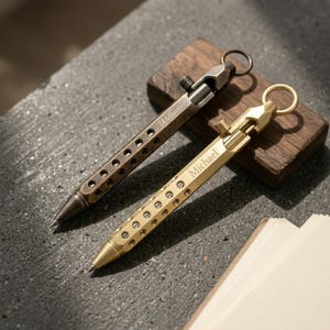 May include: Two metal pens with a perforated barrel design and a top ring attachment. One pen is finished in a bright brass tone with the name Michael engraved, while the other is a darker, aged bronze tone with the name William engraved.