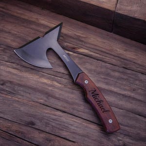 May include: A personalized axe with a brown wooden handle. The handle is engraved with the name "Michael".