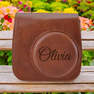 May include: Brown leather camera case with a circular flap that has the name "Olivia" engraved on it.