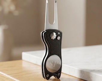 May include: A silver and black metal golf divot tool featuring a retractable two-pronged fork. The handle has a textured black grip, a circular silver inlay engraved with the letters SP, and a small silver button mechanism for deployment.