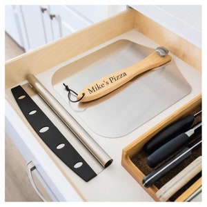 May include: A stainless steel pizza peel with a wooden handle engraved with "Mike's Pizza" rests in a drawer. A black pizza cutter and a metal pizza roller are also visible. Kitchen utensils.
