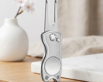 May include: A silver-toned metal golf divot repair tool featuring a two-pronged fork, a textured grip handle, and a circular centre inlay engraved with the letters JP. The tool has a sleek, metallic finish with visible screws at the base.