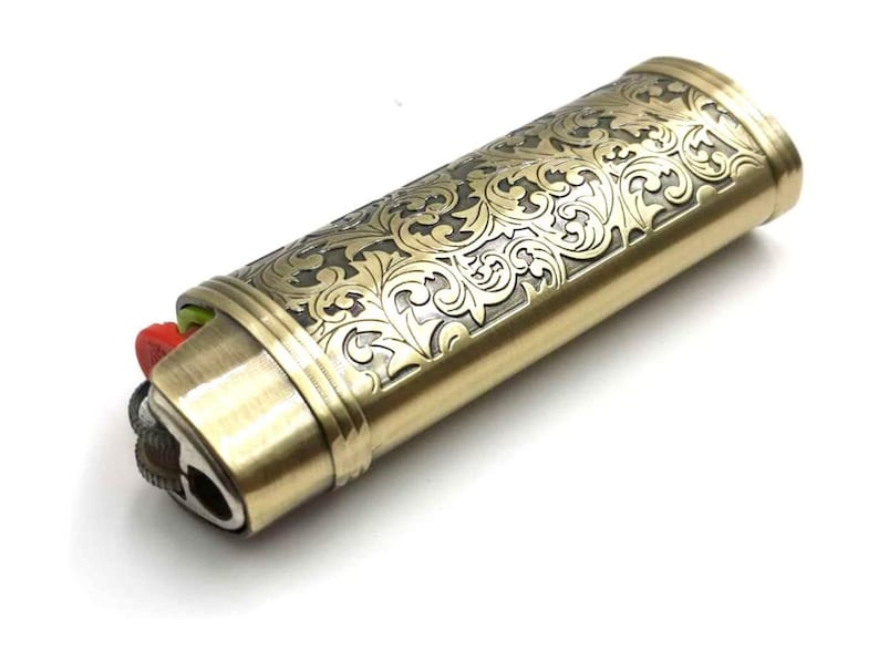 Puede incluir: Un encendedor de color dorado con un intrincado patr&oacute;n floral grabado en la superficie. El encendedor tiene un bot&oacute;n rojo y verde.