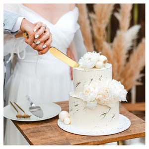 Puede incluir: Una tarta de boda blanca de dos pisos decorada con flores blancas y detalles dorados. Un cuchillo dorado para tarta corta la capa superior. Un plato con un servidor de tarta y tenedores est&aacute; sobre una mesa de madera.