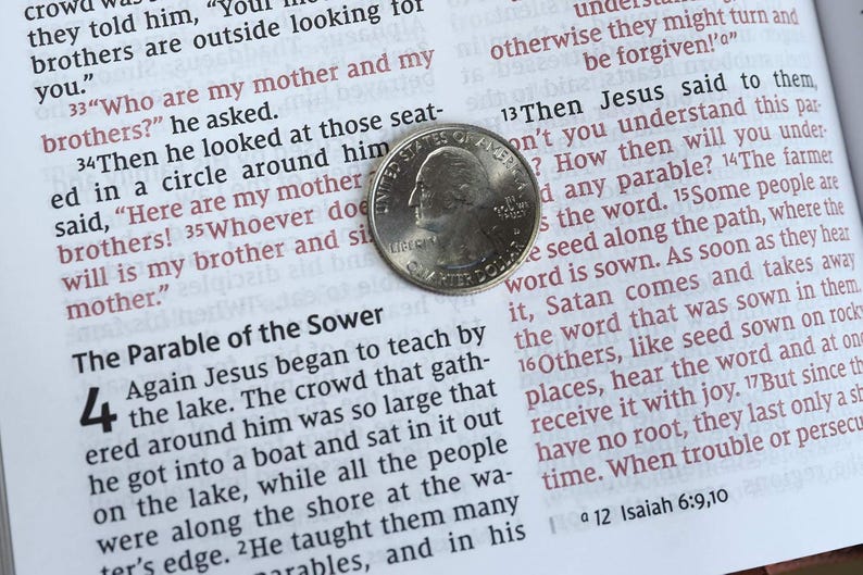 Op de afbeelding: Een zilverkleurige Amerikaanse kwartmunt rust op een open pagina van een boek met gedrukte tekst. De munt toont het profiel van George Washington en het woord Liberty, geplaatst over de tekst van de gelijkenis van de zaaier en verzen.