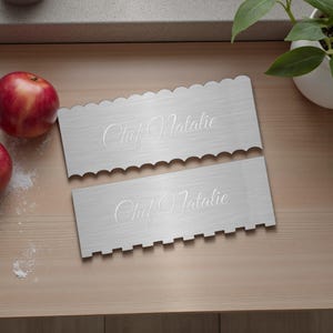 May include: Two stainless steel cake decorating tools with scalloped and notched edges. The text "Chef Natalie" is elegantly engraved on each tool. Red apples and a potted plant are in the background, suggesting a kitchen setting.