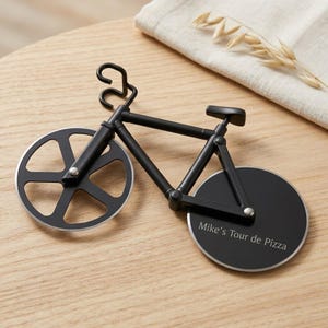 Puede incluir: Un cortador de pizza negro con forma de bicicleta, con marco y manillar. La rueda delantera tiene un diseño recortado y la rueda trasera sólida presenta el texto grabado "Mike's Tour de Pizza" en una fuente blanca sin remates.