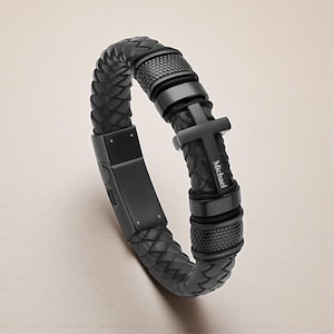 Op de afbeelding: Zwart leren gevlochten armband met een zwarte metalen kruisbedel. Op het kruis staat de naam "Michael" gegraveerd. De armband heeft een zwarte sluiting en decoratieve zwarte metalen accenten.