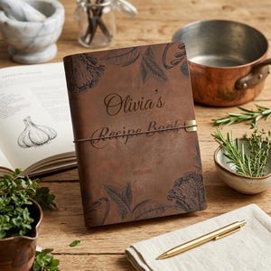Könnte beinhalten: Ein braunes Leder-Rezeptbuch mit dem Text "Olivia's Recipe Book" und floralen Illustrationen. Das Buch ist mit einem braunen Gummiband und einem goldfarbenen Verschluss verschlossen. Ein goldener Stift und eine kleine Schale mit Kräutern sind ebenfalls zu sehen.