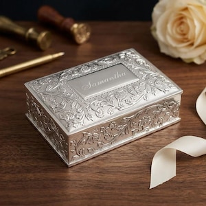 May include: A rectangular silver-toned metal box featuring an intricate embossed floral pattern across the lid and sides. The center of the lid displays a smooth, polished rectangular plaque engraved with the name Samantha in cursive script.