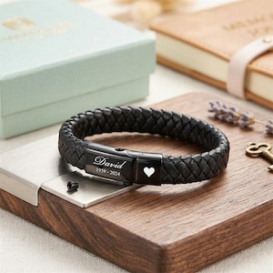 May include: Black braided leather bracelet with a stainless steel clasp. The clasp is engraved with the name "David", the years "1959-2024", and a white heart symbol. The bracelet is displayed on a wooden surface.
