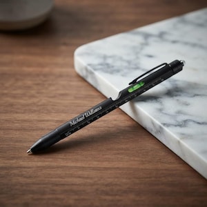 May include: A matte black multi-tool pen featuring the name Michael Williams engraved on the barrel. The pen includes a built-in ruler with inch markings, a small green spirit level, a pocket clip, and a stylus tip at the top end.