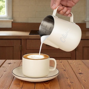 May include: A cream-colored milk frothing pitcher pouring milk into a matching coffee cup and saucer. The pitcher has the name "Olivia" printed on the side and internal measurements. The coffee has a brown crema.