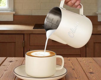 May include: A cream-colored milk frothing pitcher pouring milk into a matching coffee cup and saucer. The pitcher has the name "Olivia" printed on the side and internal measurements. The coffee has a brown crema.