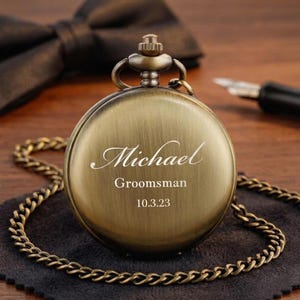 May include: Antique brass pocket watch with a chain, featuring the name "Michael", the word "Groomsman", and the date "10.3.23" engraved on the face. A black bow tie is visible in the background.