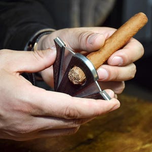 Puede incluir: Un cortapuros en V con cuerpo de madera marr&oacute;n oscuro y mecanismo de cuchilla de metal plateado. La herramienta est&aacute; colocada alrededor de un cigarro marr&oacute;n claro, mostrando la muesca en forma de V cortada en el extremo de la hoja de tabaco.